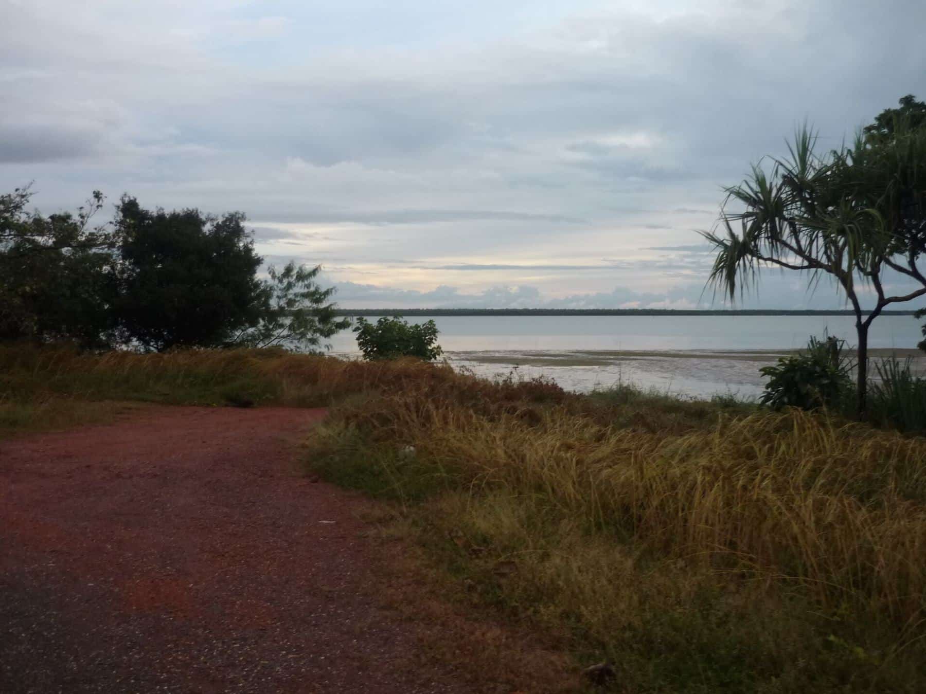 A red dirt path leading through grass and pandanus to the wide tidal Embley River near Weipa